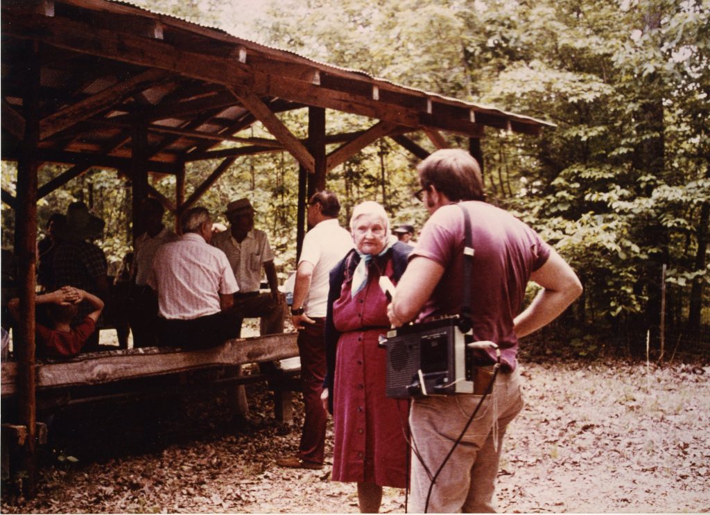 As members of the Lynn family of Shannon County, MO gather for a picnic, a documentarian speaks with the family matriarch. Photo circa 1981.