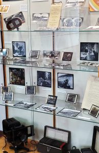 Display case with glass shelving, containing a collection of glass slides and stereograph images.