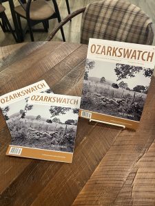 Picture of 3 copies of the newest OzarksWatch Magazine. The cover is a picture of a field in the Ozarks with stones piled at the edge of a fencerow, a wooden shed, tall grasses, and sparse trees with rolling Ozarks hills in the background.