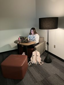 Young woman sits in "soft seating" comfortable chair with lamp on inside study room. She has her laptop on the extendable desk and is not using the footrest. 