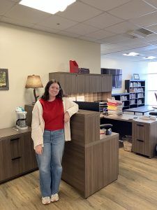 Ella Myers, a brown-haired woman, stands at her desk in blue jeans and a red shirt.