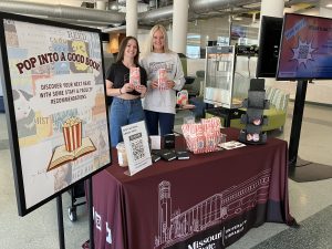 Student workers holding popcorn bags in front of a table offering popcorn, buttons, and notepads to students