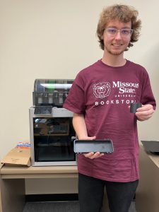 student worker standing in front of 3D printer with a case he made to hold a soldering iron