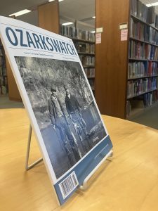 One copy of OzarksWatch magazine propped up on a book stand in a library. The magazine is on a table and bookshelves are behind it.
