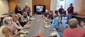 Several people gathered in a room eating and socializing at a table in front of a television screen.